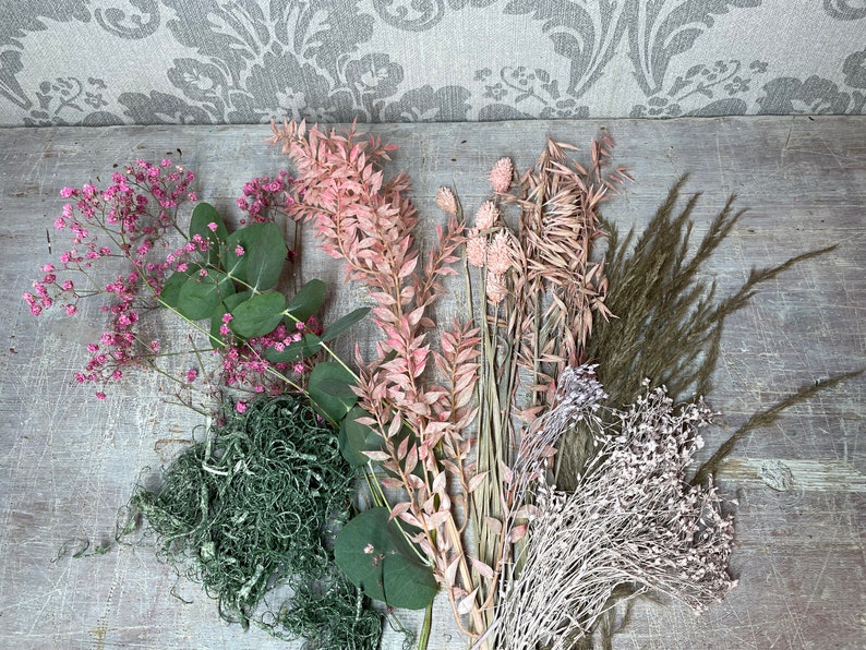 May include: An assortment of dried flowers and greenery, including pink baby's breath, eucalyptus, and feathery grasses. The arrangement features various textures and shades of pink, green, and brown, perfect for crafting or home decor.