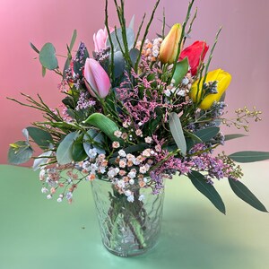 May include: A vibrant bouquet of tulips in a clear glass vase. The arrangement features pink, yellow, orange, and red tulips, accented by greenery and small white and purple flowers. The vase sits on a green surface against a pink backdrop.