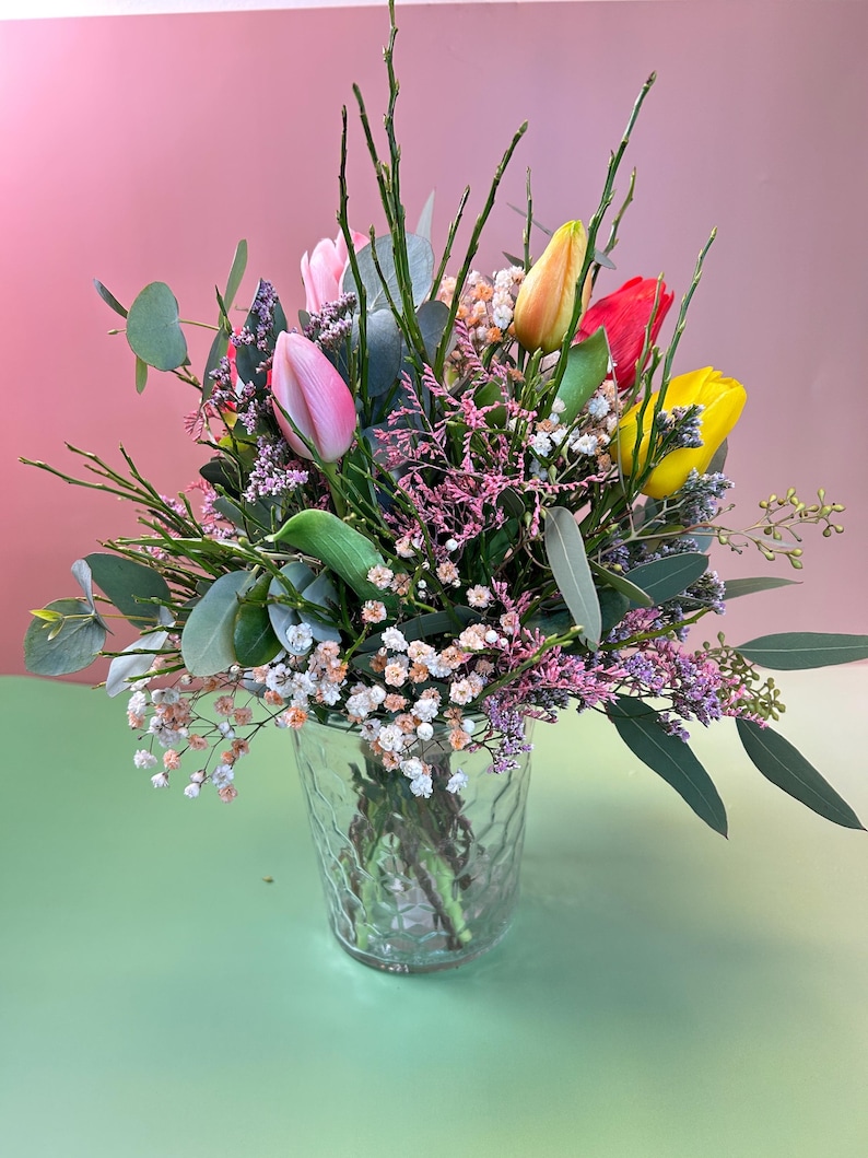 May include: A vibrant bouquet of tulips in a clear glass vase. The arrangement features pink, yellow, orange, and red tulips, accented by greenery and small white and purple flowers. The vase sits on a green surface against a pink backdrop.