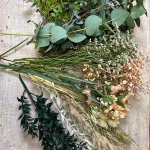 May include: An assortment of dried and fresh greenery and flowers, including eucalyptus, small white and peach-colored blooms, and various grasses. The arrangement is laid out on a light-colored wooden surface, showcasing a natural, earthy aesthetic.