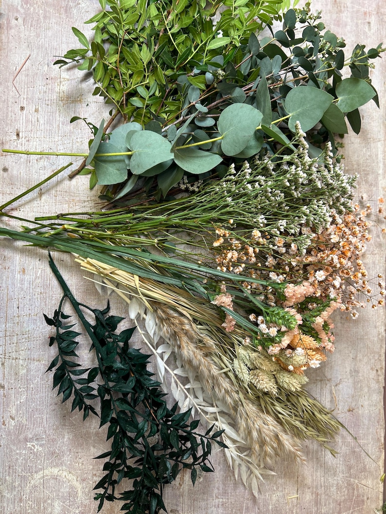 May include: An assortment of dried and fresh greenery and flowers, including eucalyptus, small white and peach-colored blooms, and various grasses. The arrangement is laid out on a light-colored wooden surface, showcasing a natural, earthy aesthetic.