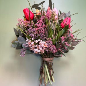 May include: A vibrant bouquet of fresh-cut flowers, featuring bright red tulips as the focal point. The arrangement includes pink and white blossoms, green foliage, and is tied with twine. The background is a soft green.