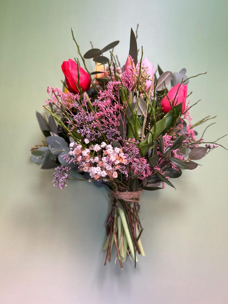 May include: A vibrant bouquet of fresh-cut flowers, featuring bright red tulips as the focal point. The arrangement includes pink and white blossoms, green foliage, and is tied with twine. The background is a soft green.