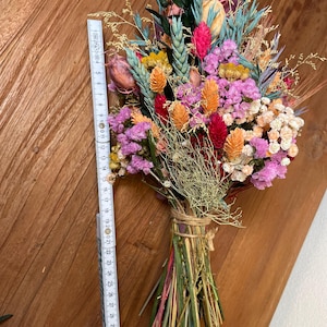 May include: A bouquet of dried flowers in shades of pink, orange, red, and green. The flowers are tied together with twine and include wheat stalks, dried grasses, and small pink and white flowers.