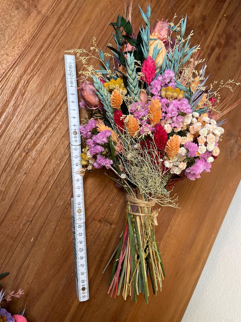 May include: A bouquet of dried flowers in shades of pink, orange, red, and green. The flowers are tied together with twine and include wheat stalks, dried grasses, and small pink and white flowers.