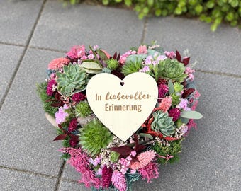 May include: A heart-shaped floral arrangement featuring green succulents, pink and magenta dried flowers, and moss. A light-colored wooden heart plaque is centered on the arrangement with the text In liebevoller Erinnerung printed in brown.