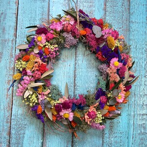 May include: A colourful circular wreath made from dried flowers in shades of pink, yellow, orange, and purple. The wreath hangs against a weathered, turquoise-painted wooden background, highlighting its natural textures and varied hues.