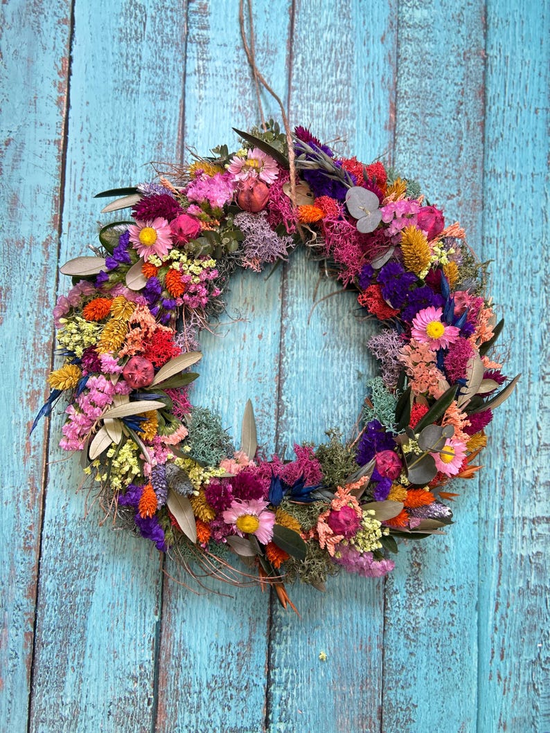 May include: A colourful circular wreath made from dried flowers in shades of pink, yellow, orange, and purple. The wreath hangs against a weathered, turquoise-painted wooden background, highlighting its natural textures and varied hues.