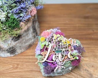 May include: A heart-shaped floral arrangement with the words "with love" in a wooden script. The arrangement features a variety of colorful flowers and moss. A second arrangement with purple flowers is in the background.