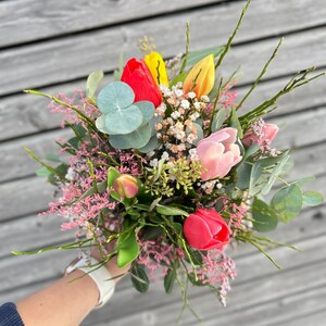 May include: A vibrant bouquet featuring red, yellow, and pink tulips, accented with sprigs of baby's breath, eucalyptus leaves, and delicate pink flowers. The arrangement is held in a hand, showcasing a fresh and colorful floral display.