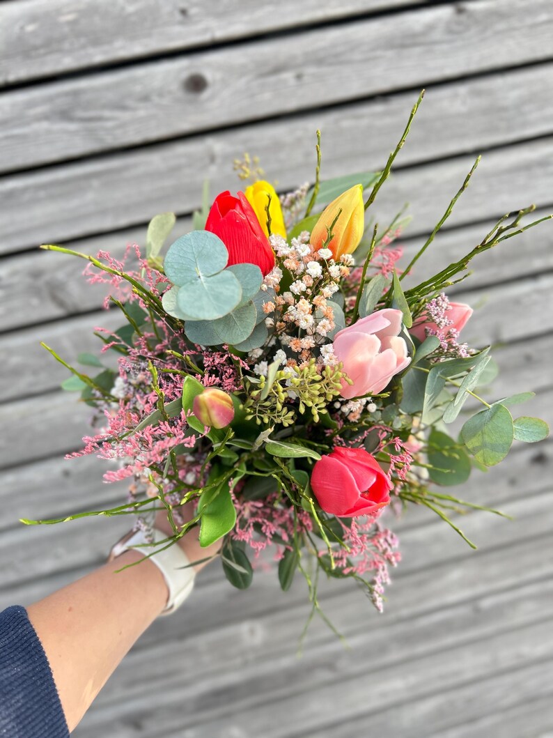 May include: A vibrant bouquet featuring red, yellow, and pink tulips, accented with sprigs of baby's breath, eucalyptus leaves, and delicate pink flowers. The arrangement is held in a hand, showcasing a fresh and colorful floral display.