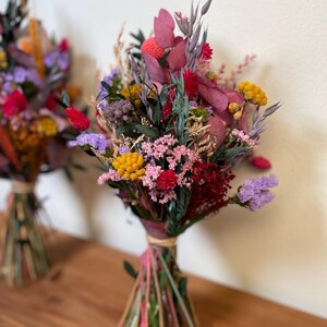 May include: A bouquet of dried flowers in shades of pink, purple, yellow, and red. The flowers are tied together with a natural twine and are arranged in a rustic style.