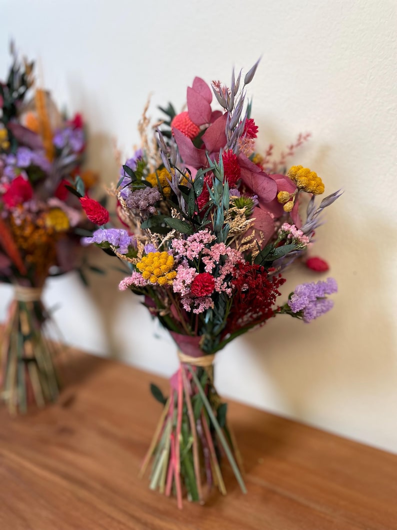 May include: A bouquet of dried flowers in shades of pink, purple, yellow, and red. The flowers are tied together with a natural twine and are arranged in a rustic style.