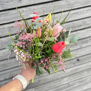 May include: A vibrant bouquet of fresh-cut flowers, featuring bright red, pink, and yellow tulips, accented with green foliage and delicate pink filler flowers. The arrangement is held in a hand, showcasing the natural beauty of the floral composition.