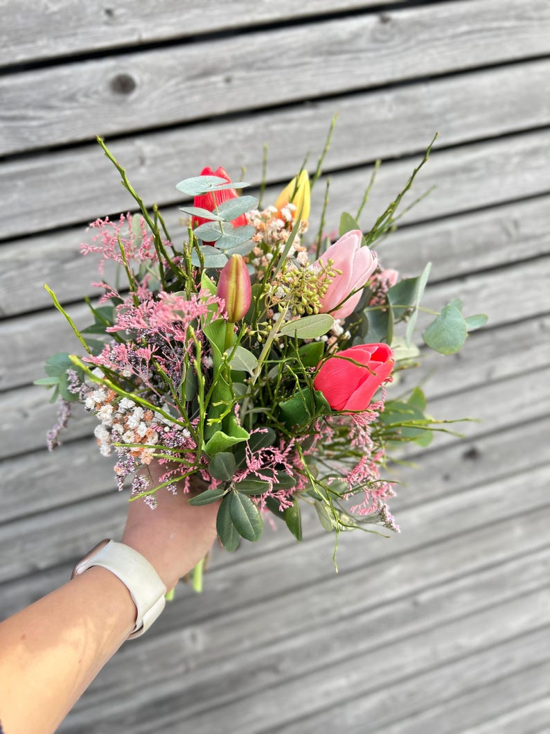May include: A vibrant bouquet of fresh-cut flowers, featuring bright red, pink, and yellow tulips, accented with green foliage and delicate pink filler flowers. The arrangement is held in a hand, showcasing the natural beauty of the floral composition.