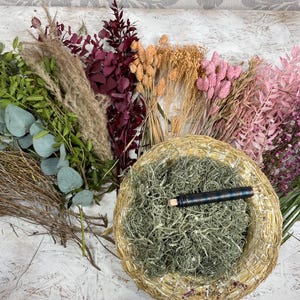 May include: An assortment of dried flowers and greenery, including eucalyptus, pampas grass, and various pink and purple blooms. A woven basket filled with green moss and a dark pen is in the center. The arrangement is on a white surface.