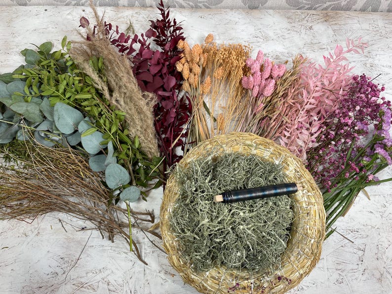 May include: An assortment of dried flowers and greenery, including eucalyptus, pampas grass, and various pink and purple blooms. A woven basket filled with green moss and a dark pen is in the center. The arrangement is on a white surface.