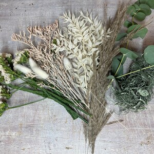 May include: An assortment of dried flowers and greenery, including pampas grass, eucalyptus, and various dried blooms in shades of white, beige, and green. The arrangement is laid out on a light-colored wooden surface.