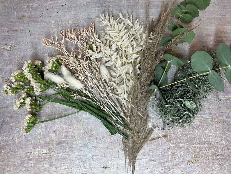 May include: An assortment of dried flowers and greenery, including pampas grass, eucalyptus, and various dried blooms in shades of white, beige, and green. The arrangement is laid out on a light-colored wooden surface.