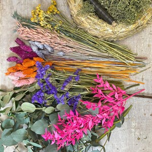 May include: A collection of dried flowers and greenery, including pink, purple, orange, and yellow blooms, along with eucalyptus leaves. A woven wreath base with green moss and a dark brown spool of wire is also present. The arrangement is set on a light wood surface.