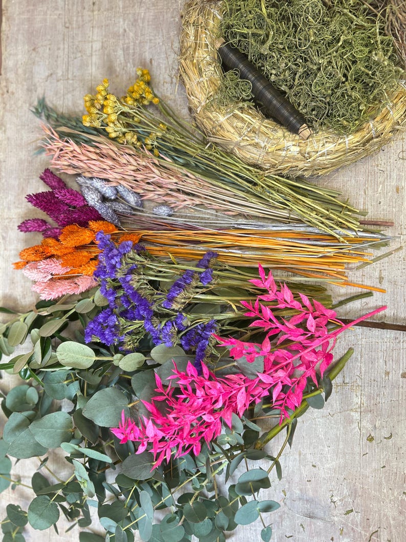 May include: A collection of dried flowers and greenery, including pink, purple, orange, and yellow blooms, along with eucalyptus leaves. A woven wreath base with green moss and a dark brown spool of wire is also present. The arrangement is set on a light wood surface.