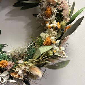 May include: A handmade wreath with a variety of dried flowers and foliage. The wreath features olive leaves, orange and white flowers, and fluffy beige accents. The wreath is set against a plain white background.