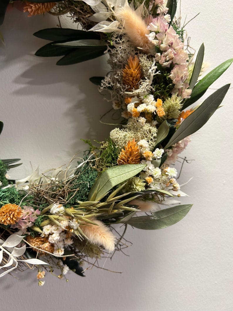 May include: A handmade wreath with a variety of dried flowers and foliage. The wreath features olive leaves, orange and white flowers, and fluffy beige accents. The wreath is set against a plain white background.