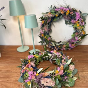 May include: Two colourful floral wreaths made from dried flowers and foliage, displayed on a wooden surface. Two mint green lamps are visible in the background. The wreaths feature pink, yellow, and green elements.