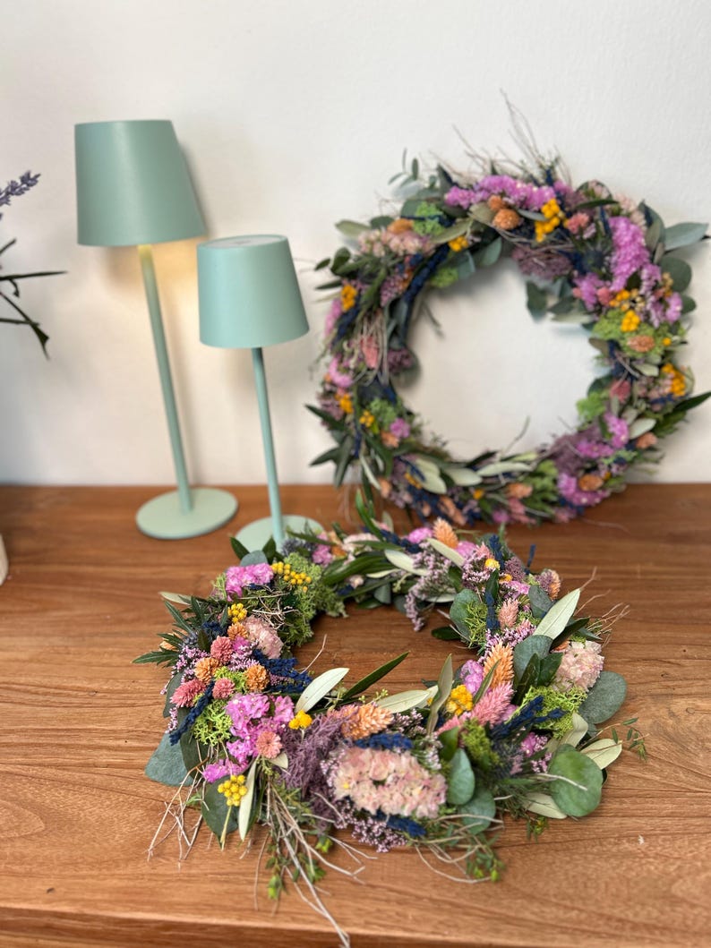 May include: Two colourful floral wreaths made from dried flowers and foliage, displayed on a wooden surface. Two mint green lamps are visible in the background. The wreaths feature pink, yellow, and green elements.