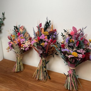 May include: Three bouquets of dried flowers in shades of pink, purple, yellow, and orange. The bouquets are tied with twine and have a rustic, natural look.