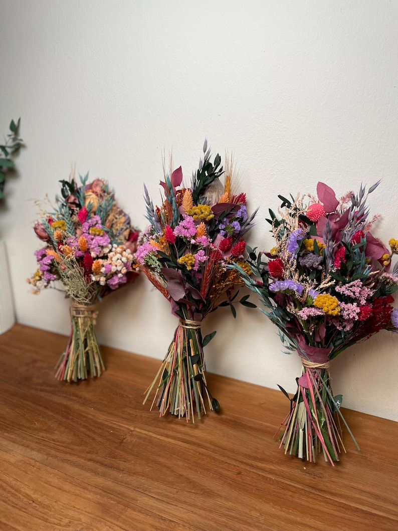 May include: Three bouquets of dried flowers in shades of pink, purple, yellow, and orange. The bouquets are tied with twine and have a rustic, natural look.