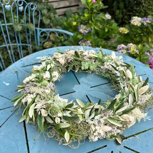 May include: A decorative circular wreath made from dried flowers and foliage, including olive leaves and white flowers. The wreath is displayed on a light blue table, with a garden setting in the background.