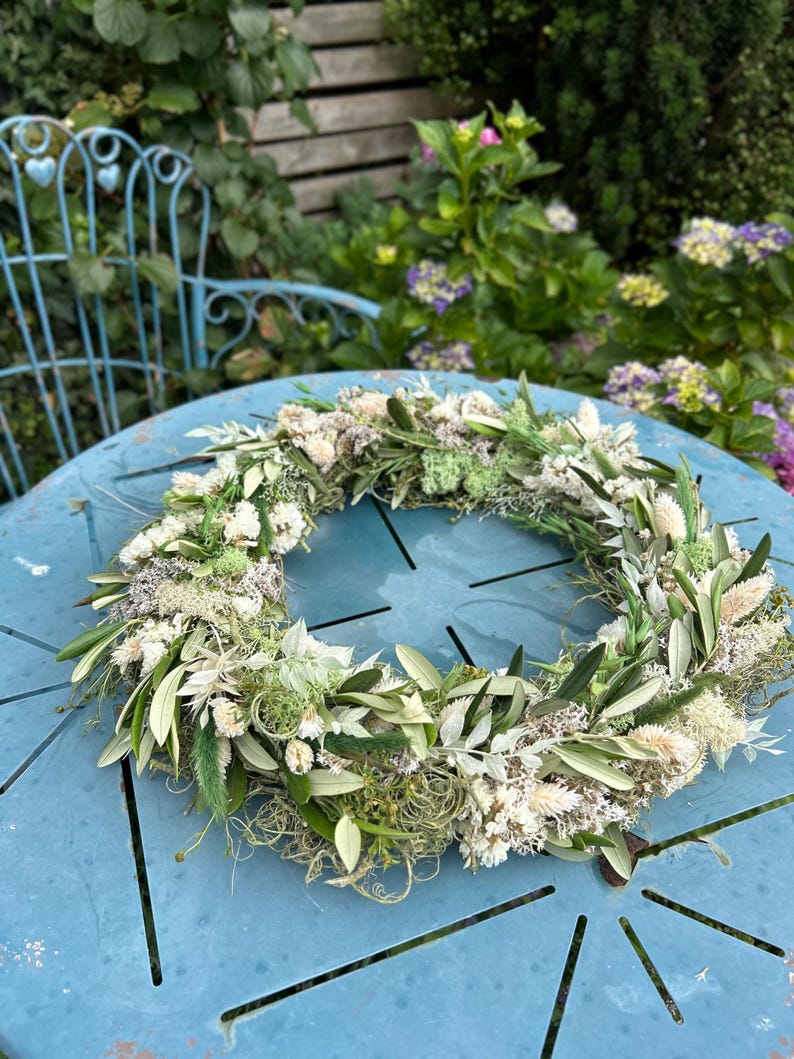 May include: A decorative circular wreath made from dried flowers and foliage, including olive leaves and white flowers. The wreath is displayed on a light blue table, with a garden setting in the background.