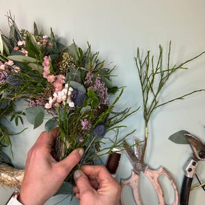 May include: A floral arrangement being assembled, featuring a wreath base with pink, purple, and green foliage. The hands are working with the flowers, using scissors and twine. The scene suggests a craft project or floral design.