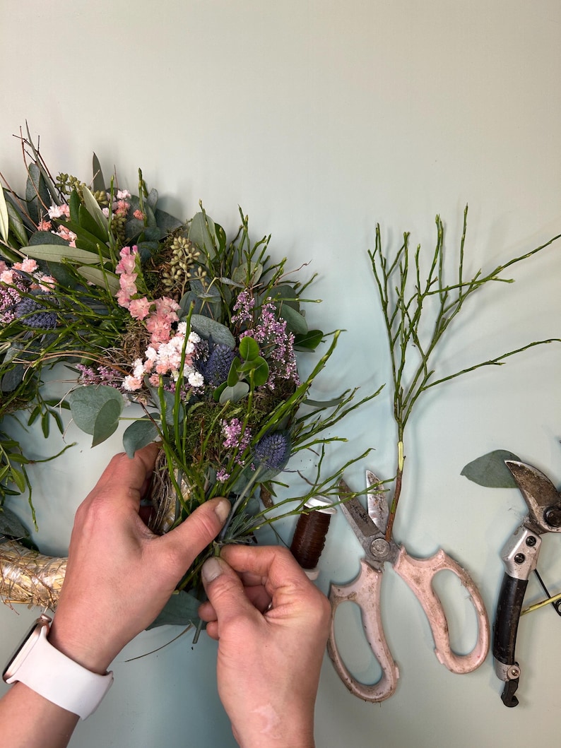 May include: A floral arrangement being assembled, featuring a wreath base with pink, purple, and green foliage. The hands are working with the flowers, using scissors and twine. The scene suggests a craft project or floral design.