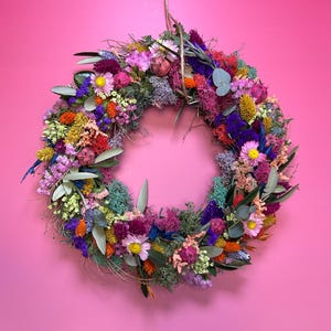 May include: A colourful, circular floral wreath made from dried flowers and foliage. The wreath, suspended by twine, features pink, purple, yellow, orange, and blue flowers. It is set against a bright pink wall, creating a striking contrast.
