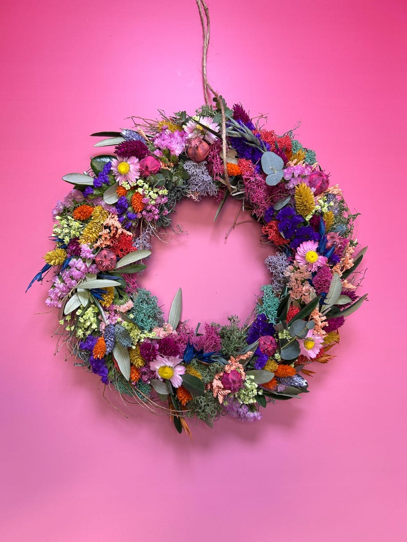 May include: A colourful, circular floral wreath made from dried flowers and foliage. The wreath, suspended by twine, features pink, purple, yellow, orange, and blue flowers. It is set against a bright pink wall, creating a striking contrast.