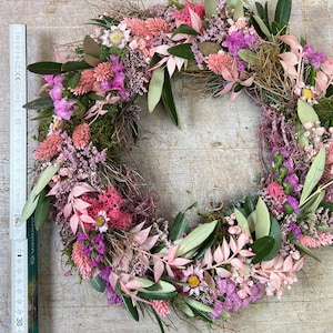 May include: A circular floral wreath made from dried flowers and foliage. The wreath includes pink, purple, and green elements, such as leaves, small flowers, and moss. The wreath's diameter is approximately 30 cm.