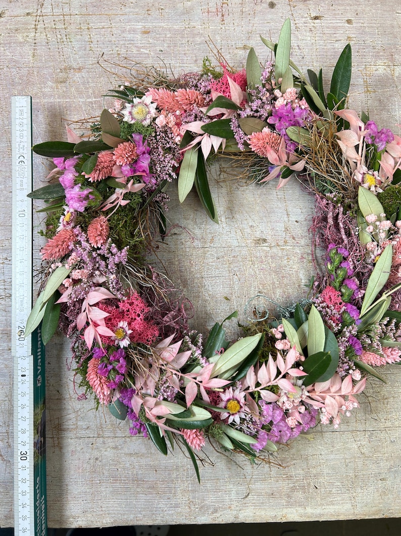 May include: A circular floral wreath made from dried flowers and foliage. The wreath includes pink, purple, and green elements, such as leaves, small flowers, and moss. The wreath's diameter is approximately 30 cm.