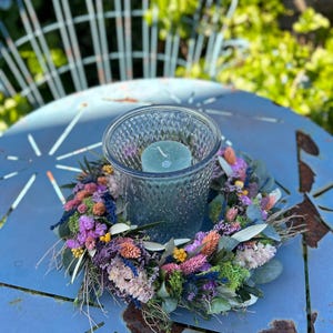 May include: A teal glass candle holder with a teal candle sits on a colourful dried flower wreath. The wreath features purple, pink, orange, and green flowers. The arrangement rests on a distressed blue metal table.