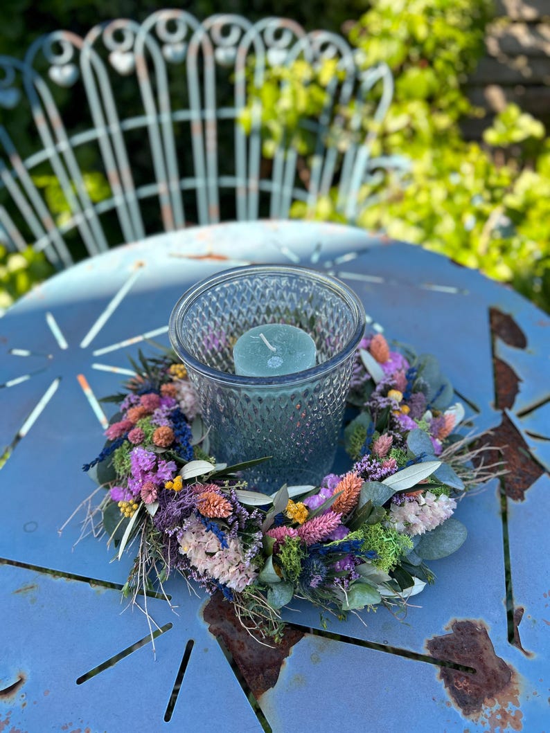 May include: A teal glass candle holder with a teal candle sits on a colourful dried flower wreath. The wreath features purple, pink, orange, and green flowers. The arrangement rests on a distressed blue metal table.