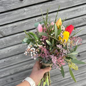 May include: A colorful bouquet of fresh flowers, including pink, yellow, and red tulips, with green foliage and small white and pink blossoms. The bouquet is tied with twine and held in a person's hand.