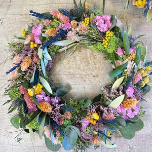 May include: A colourful floral wreath composed of dried flowers in pink, yellow, blue, and orange, with green foliage. The circular wreath is ideal for decorating a home, with a diameter of approximately 30 cm.