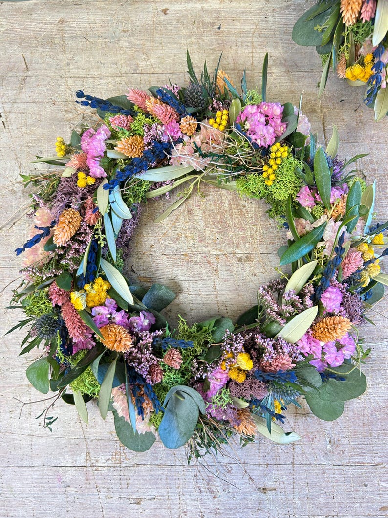May include: A colourful floral wreath composed of dried flowers in pink, yellow, blue, and orange, with green foliage. The circular wreath is ideal for decorating a home, with a diameter of approximately 30 cm.