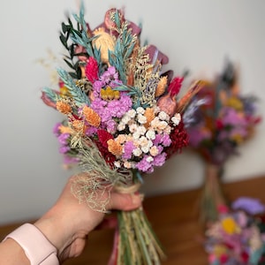 May include: A bouquet of dried flowers in shades of pink, orange, and red. The bouquet is tied with twine and includes various types of dried flowers, including baby's breath, statice, and strawflower.