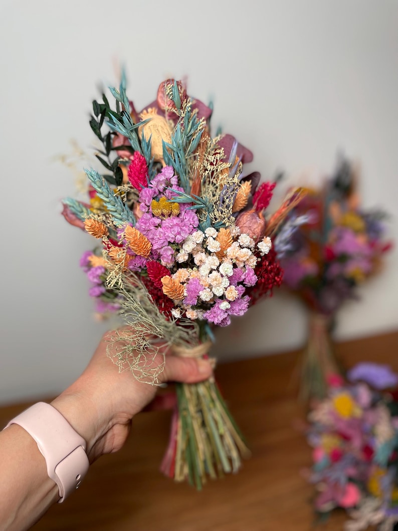May include: A bouquet of dried flowers in shades of pink, orange, and red. The bouquet is tied with twine and includes various types of dried flowers, including baby's breath, statice, and strawflower.
