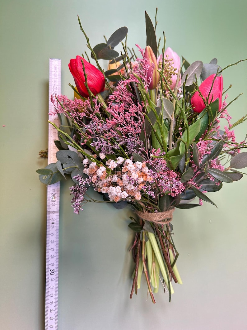 May include: A vibrant bouquet of flowers, featuring red tulips, pink and white filler flowers, and green foliage. The stems are tied with twine. The bouquet is approximately 12 inches long.