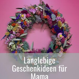 May include: A vibrant floral wreath composed of dried flowers in shades of pink, purple, yellow, and green, set against a pink backdrop. The image includes text in German: "Langlebige Geschenkideen f&uuml;r Mama".