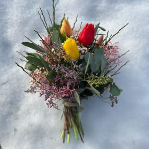 May include: A vibrant bouquet of fresh-cut flowers, featuring three tulips in yellow, red, and orange hues. The arrangement includes greenery, pink and purple filler flowers, and is tied with twine. The bouquet rests on a snowy surface.