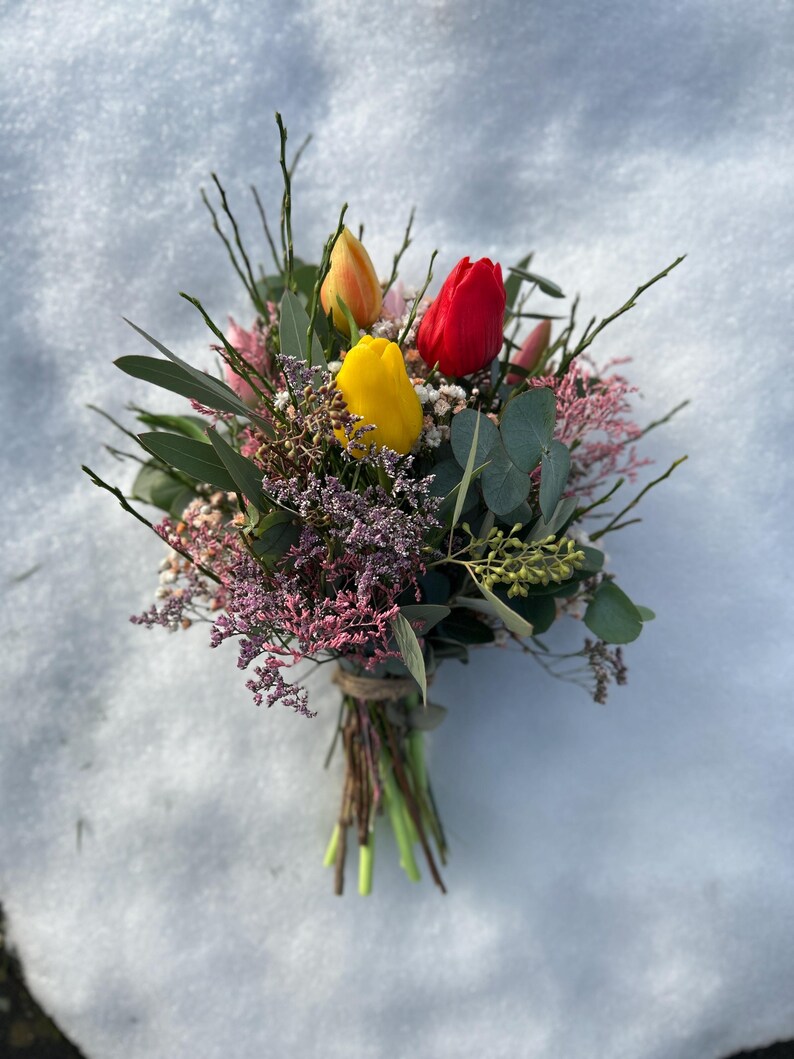 May include: A vibrant bouquet of fresh-cut flowers, featuring three tulips in yellow, red, and orange hues. The arrangement includes greenery, pink and purple filler flowers, and is tied with twine. The bouquet rests on a snowy surface.